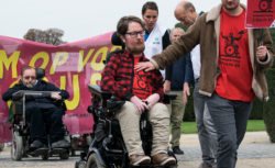 Photograph of activists marching towards the camera. In the foreground, a wheelchair user is starting to manoeuvre down a slope. A person next to him has placed their right hand on the wheelchair user's chest. In their left hand, they hold a placard but the writing is not identifiable. Both the wheelchair user and the person next to them have identical T-shirts. The T-shirts are red with a black outline of a wheelchair user. In the background are several activists. They are sitting and standing in front of a large purple banner, but the writing is obscured due to the position of the activists.