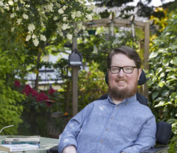 Miro sits in a black chair, facing the camera, and is smiling. He has short brown hair, rectangular, brown, thick rimmed glasses, and a short beard. He is wearing a blue shirt. There is a garden table next to him, which is painted green. There are books on the table authored by Foucault and Chomsky. In the background there are flowers and garden bushes 